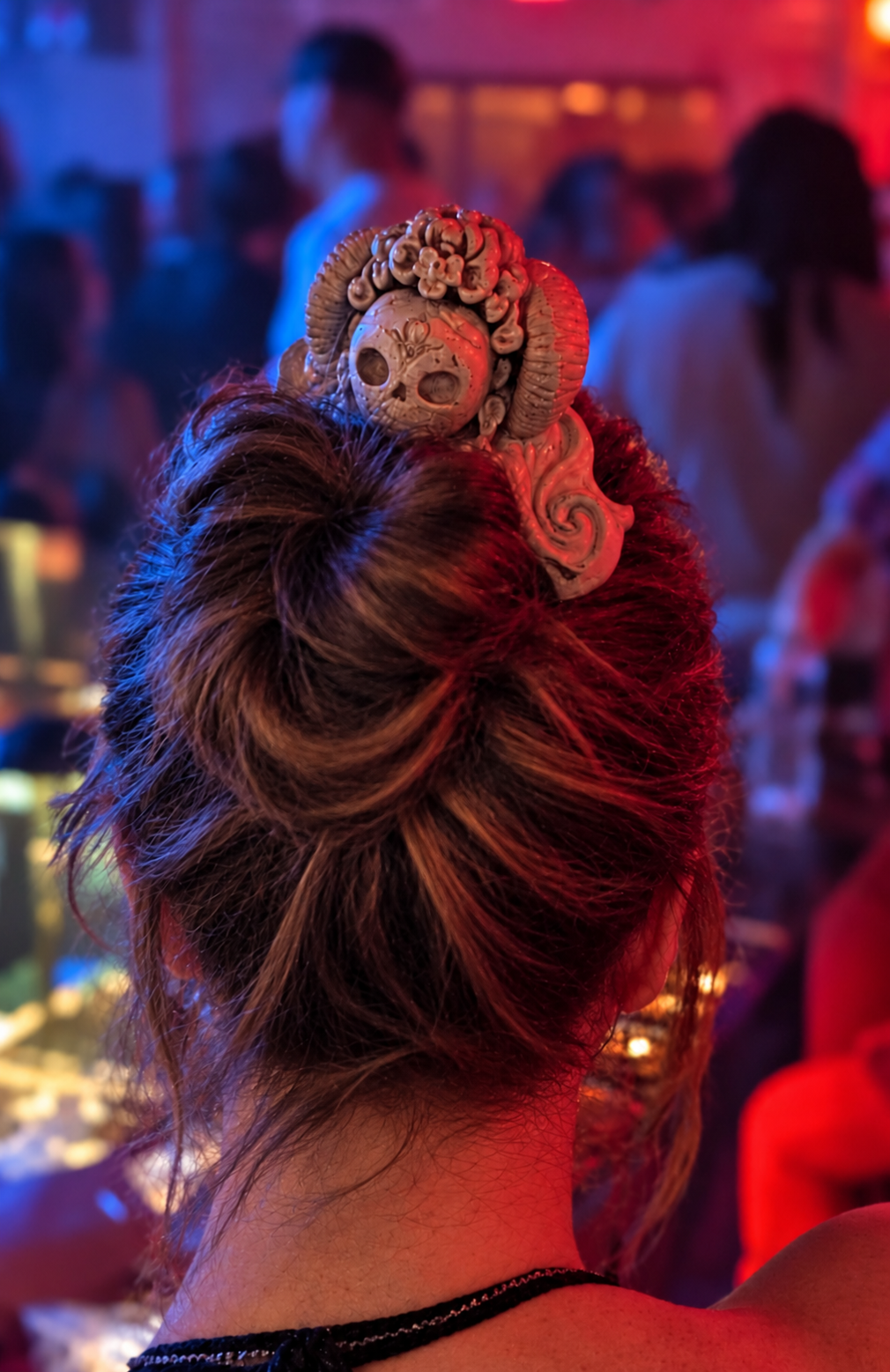 Close-up of the ivory skull and rose comb in Maya's highlighted bun under red and blue club lights.